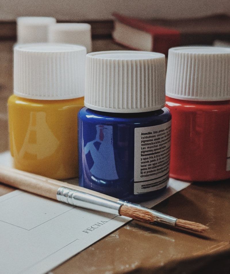 Three fabric paint jars in yellow, blue, and red with a paintbrush on a wooden surface.